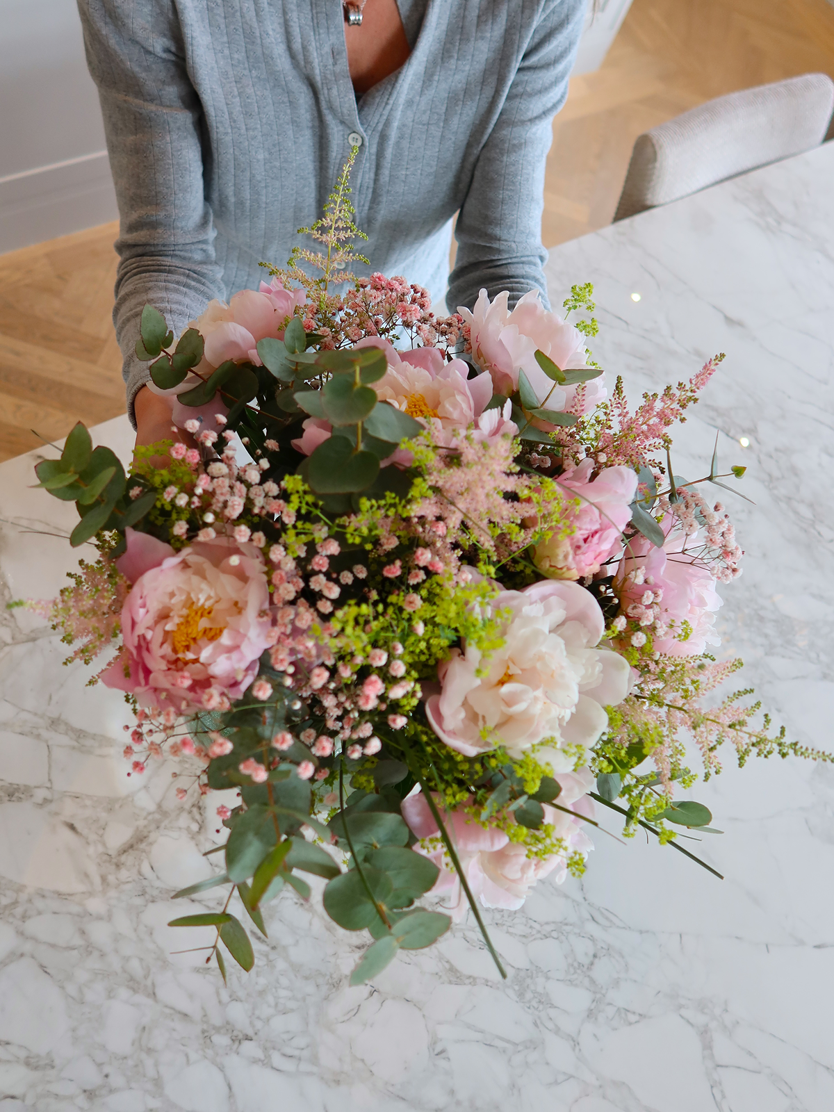 Nærbilde ovenfra av en stor bukett med lyserosa peoner, små rosa blomster, grønt og eucalyptus holdt over et marmorbord.