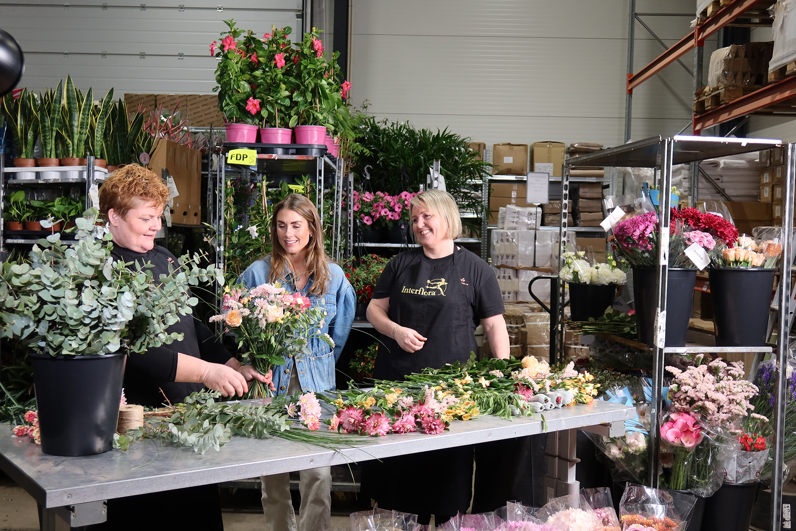 Tre kvinner står bak et arbeidsbord i et blomsterlager. Kvinnen i midten holder en bukett i rosa, fersken og grønt.
