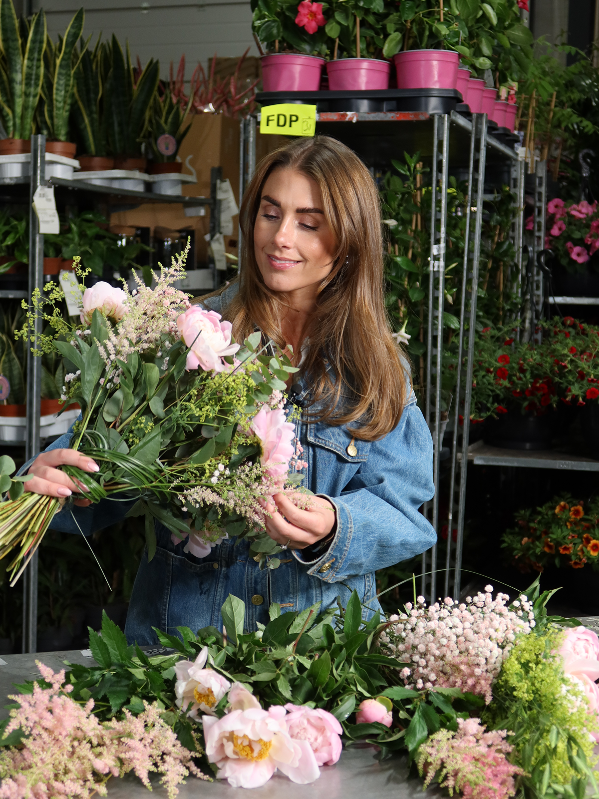 En kvinne holder en luftig bukett med lyserosa peoner, grønt og små rosa blomster over et bord med avskårne blomster.
